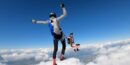 Two people are skydiving above a cloudscape, with one leading in a grey jumpsuit and another floating behind, wearing a pink jumpsuit and a harness.
