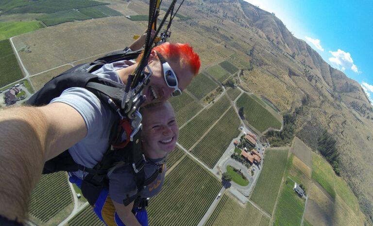 Two people are tandem skydiving, smiling with a selfie stick. They are high above a patchwork of fields with a mountainous backdrop.