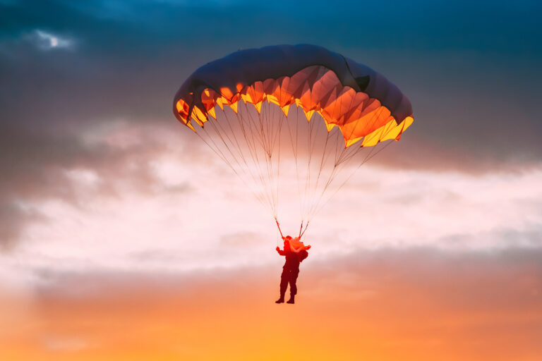 A person is parachuting against a vibrant sunset sky with shades of orange and blue. The parachute is illuminated from within, creating a striking contrast.